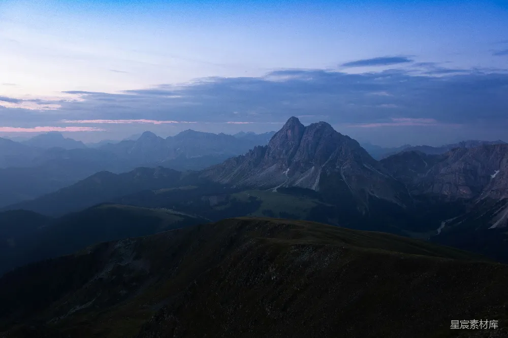这张图片展示了一幅宁静的山景，日落时分的天空呈现出柔和的色彩，与远处山脉的轮廓形成鲜明对比。山峦在暮色中显得格外壮丽，给人一种宁静与平和的感觉。适合用作户外旅行、自然摄影或宁静生活的背景素材。