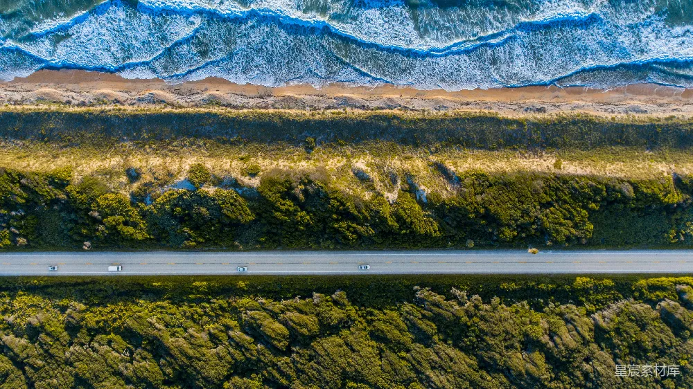 这张图片展示了从高空俯瞰的海岸公路场景，公路两旁是郁郁葱葱的植被和沙质海滩，远处是波涛汹涌的大海，画面中还有几辆行驶的汽车。整体风格清新自然，适合用于展示旅行、自然风光或旅行目的地的宣传。
