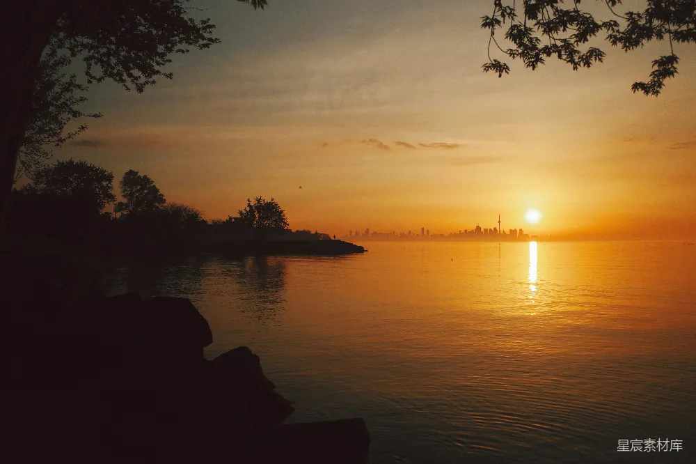 这张图片捕捉到了城市日落的宁静时刻。夕阳的余晖洒在地平线的另一边，照亮了城市的天际线。水面反射着金色的光芒，与天空的暖色调相映成趣。前景中，树木和岩石的剪影为画面增添了深度和对比。整个场景充满了宁静与美丽，适合用于城市风光和自然摄影的背景。