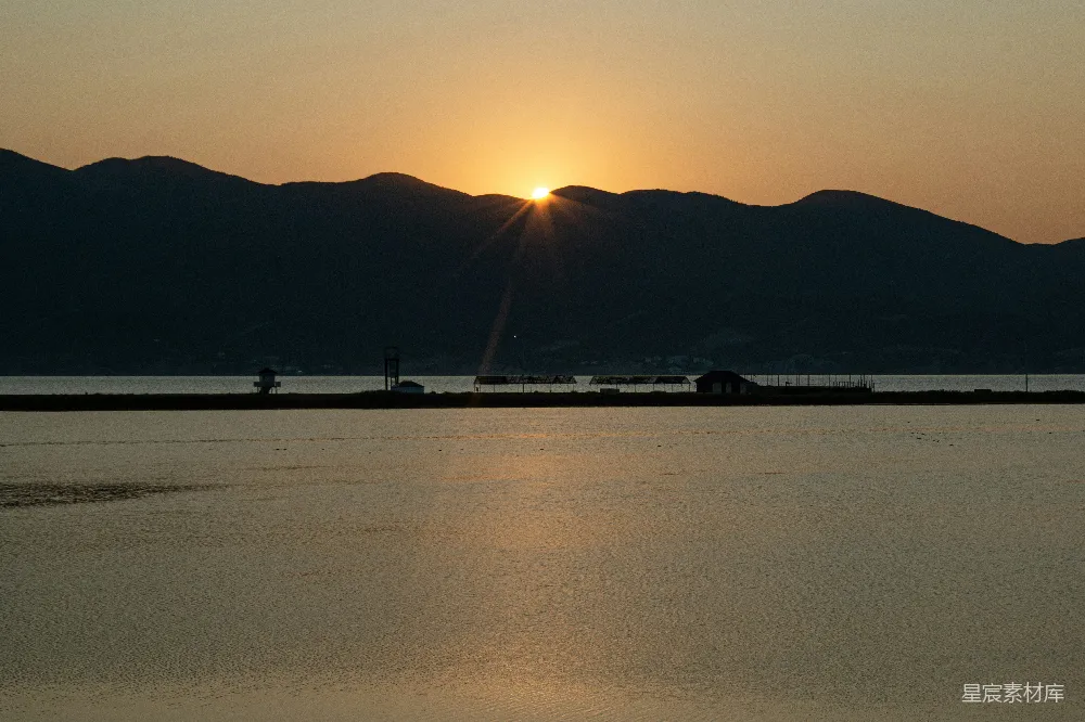 图片展示了一个宁静的湖畔景象，夕阳正缓缓落下，金色的光芒洒在湖面上，远处的山峦在暮色中显得轮廓分明。湖边的建筑在夕阳的映衬下显得格外宁静与祥和。整个场景展现了自然的美丽与宁静。