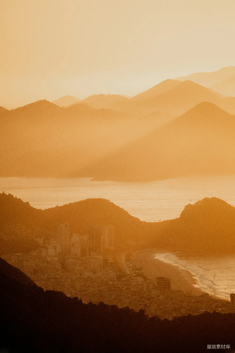 这张图片展示了一个宁静而美丽的夕阳场景。画面中，金色的阳光洒在连绵起伏的山脉上，形成了一层温暖的滤镜效果。在山的脚下一侧，可以看到一座城市的轮廓，建筑物和海滩在落日的余晖中显得格外宁静。整个画面给人一种平和而壮丽的感觉，适合用作旅行、自然或