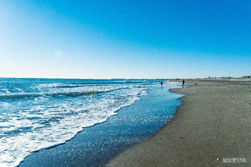 这张图片展示了海滩上轻柔的海浪拍打着沙滩，天空湛蓝，远处有几个人在享受这片宁静的海岸。适合用作旅行、自然风光或休闲放松的背景图片。