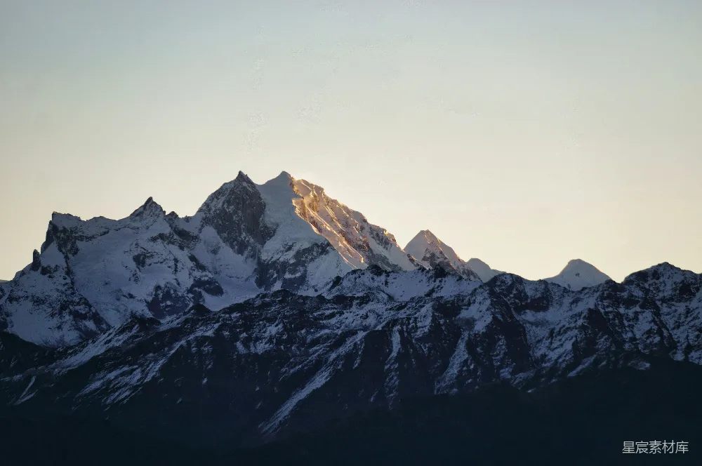 这张图片展示了日出时分的雪山景象，阳光从山峰的侧面照射下来，形成一道金色的光芒，照亮了山峰的轮廓，背景是渐变的天色，给人一种宁静而壮丽的感觉。适合用于自然风光、户外探险或清晨主题的场景。