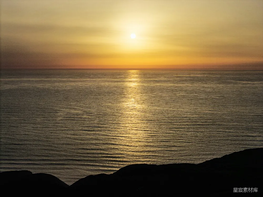 夕阳缓缓落下，金色的光芒洒在平静的海面上，形成一条美丽的光线。远处的天边被染上了橙黄色的色彩，近处的岩石在夕阳的余晖中显得更加黑暗。整个画面给人一种宁静而祥和的感觉，适合用作自然风光或旅行摄影的素材。