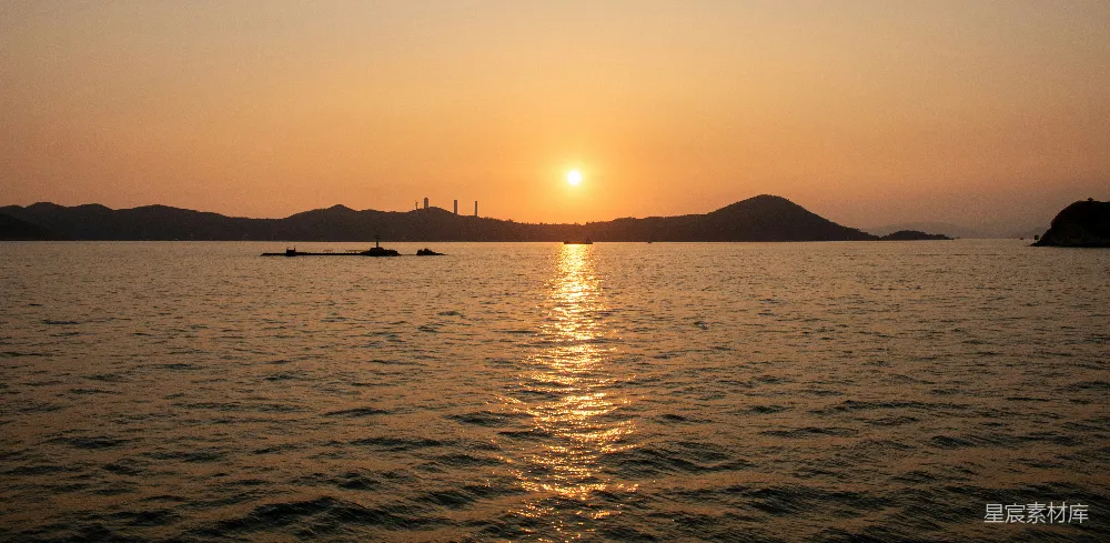 这张图片展示了一个宁静的海港在夕阳下的美景。太阳正在地平线上缓缓落下，金色的光芒洒在海面上，形成一道耀眼的光芒。远处的山脉和海港的轮廓在日落的余晖中显得格外宁静。这张图片适合用于表现自然风光、日落美景以及宁静的氛围。