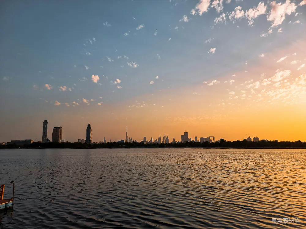 这张图片展示了城市湖边的日落美景。湖面上波光粼粼，远处的城市天际线在夕阳的映照下显得宁静而美丽。天空中散布着几朵白云，为画面增添了层次感。这张图片适合用作旅游宣传、城市风光展示或个人写真集的封面。
