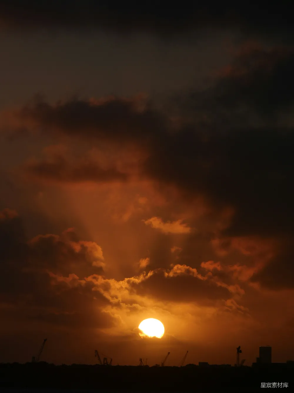 这张图片捕捉到了夕阳在云层中缓缓下沉的壮丽时刻。夕阳的余晖洒在工业区的轮廓上，形成了鲜明的对比。云层的橙黄色与地平线下的建筑形成了一幅宁静而神秘的景象。这不仅展现了自然与人类活动的和谐共存，也突显了城市黄昏时分的独特美感。