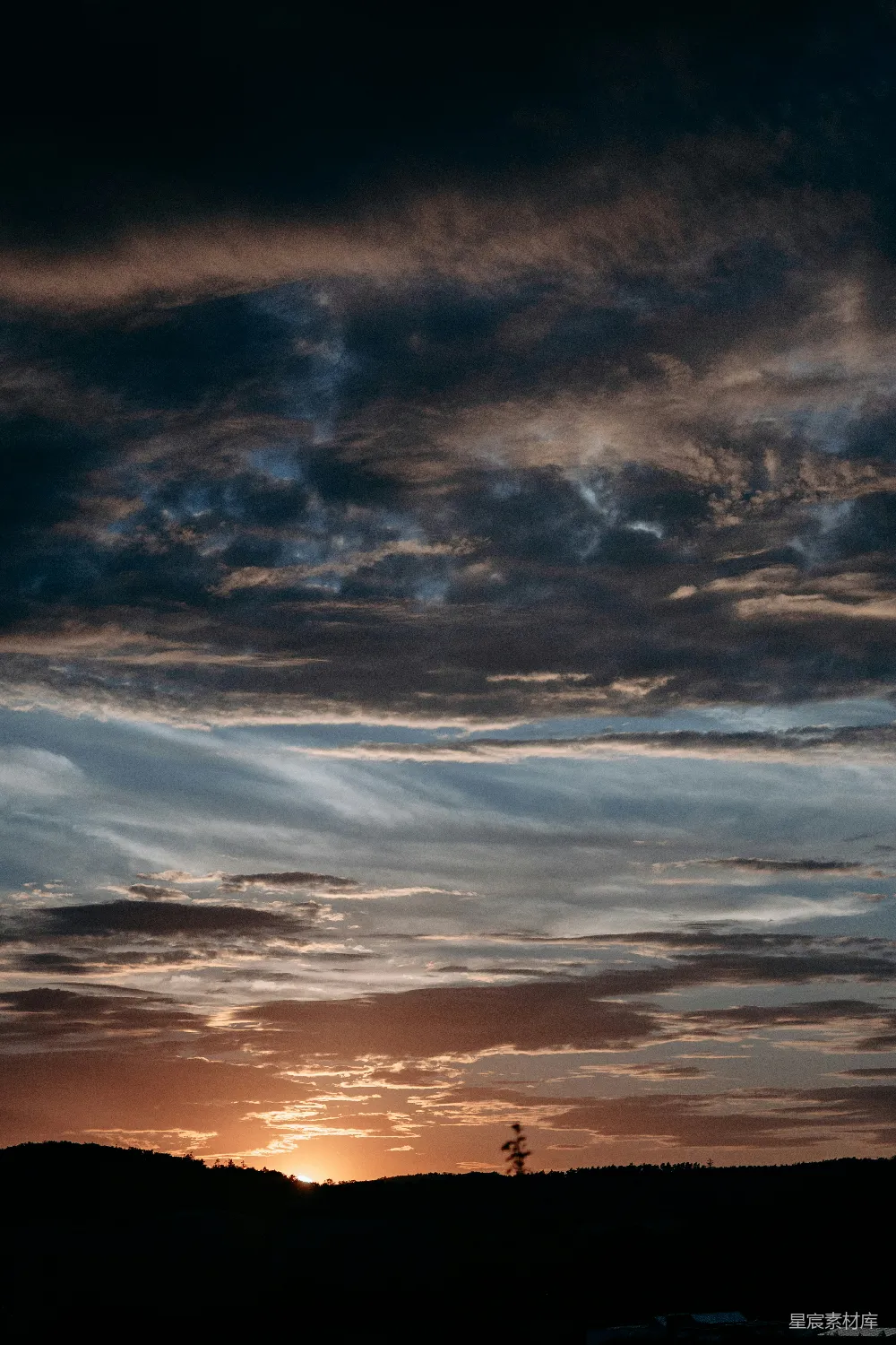 图片展示了一幅宁静的山景，夕阳的余晖洒在云层和山峦上，营造出一种温暖而宁静的氛围。天空中的云彩层次分明，色彩丰富，从深蓝到橙色，展现了自然的壮丽与变幻。山脚下有一棵树的剪影，增添了几分生动与立体感。这幅图适合用作自然风光的背景，或是表达宁静