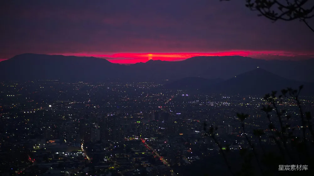这张图片展示了城市夜景与日落的美丽景象。城市灯火辉煌，与远处的山脉和红色的日落形成鲜明对比，营造出一种宁静而美丽的氛围。适合用于城市风光、旅游宣传或自然与城市结合的主题。