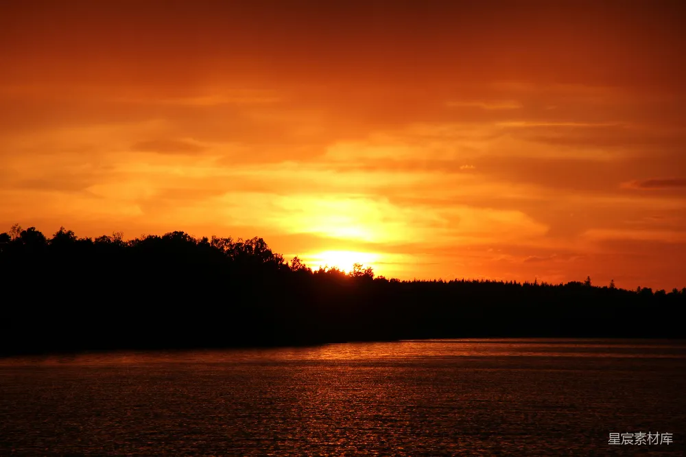 这张图片展示了一幅宁静的湖畔夕阳景色。天空被夕阳染成了橙黄色，湖面反射着温暖的光芒。远处的树梢在夕阳的映照下形成剪影，整个场景显得非常宁静和美丽，适合用来表达宁静、自然或日落等主题。