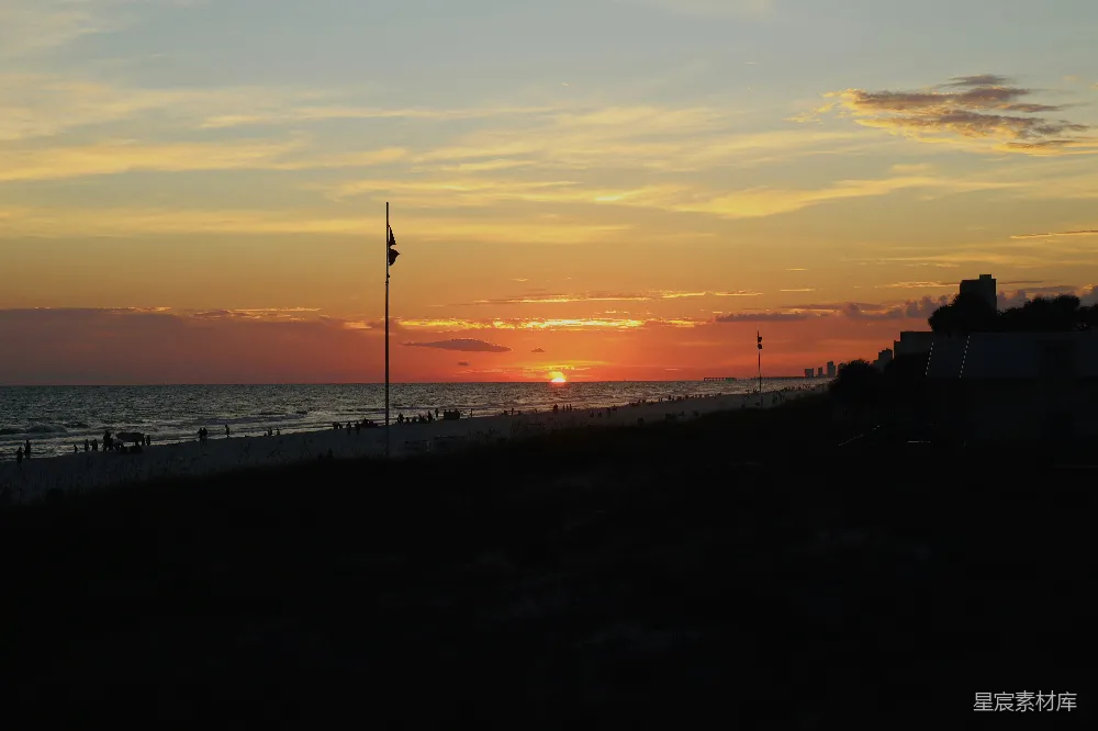 这张图片展示了海滩日落时的宁静美景。夕阳的余晖洒在海面上，天空被染成了橙红色，海浪轻轻拍打着沙滩。远处的建筑和旗帜在夕阳的余晖中形成了剪影，增添了画面的层次感。适合用作自然风光、日落、宁静氛围的背景图片。