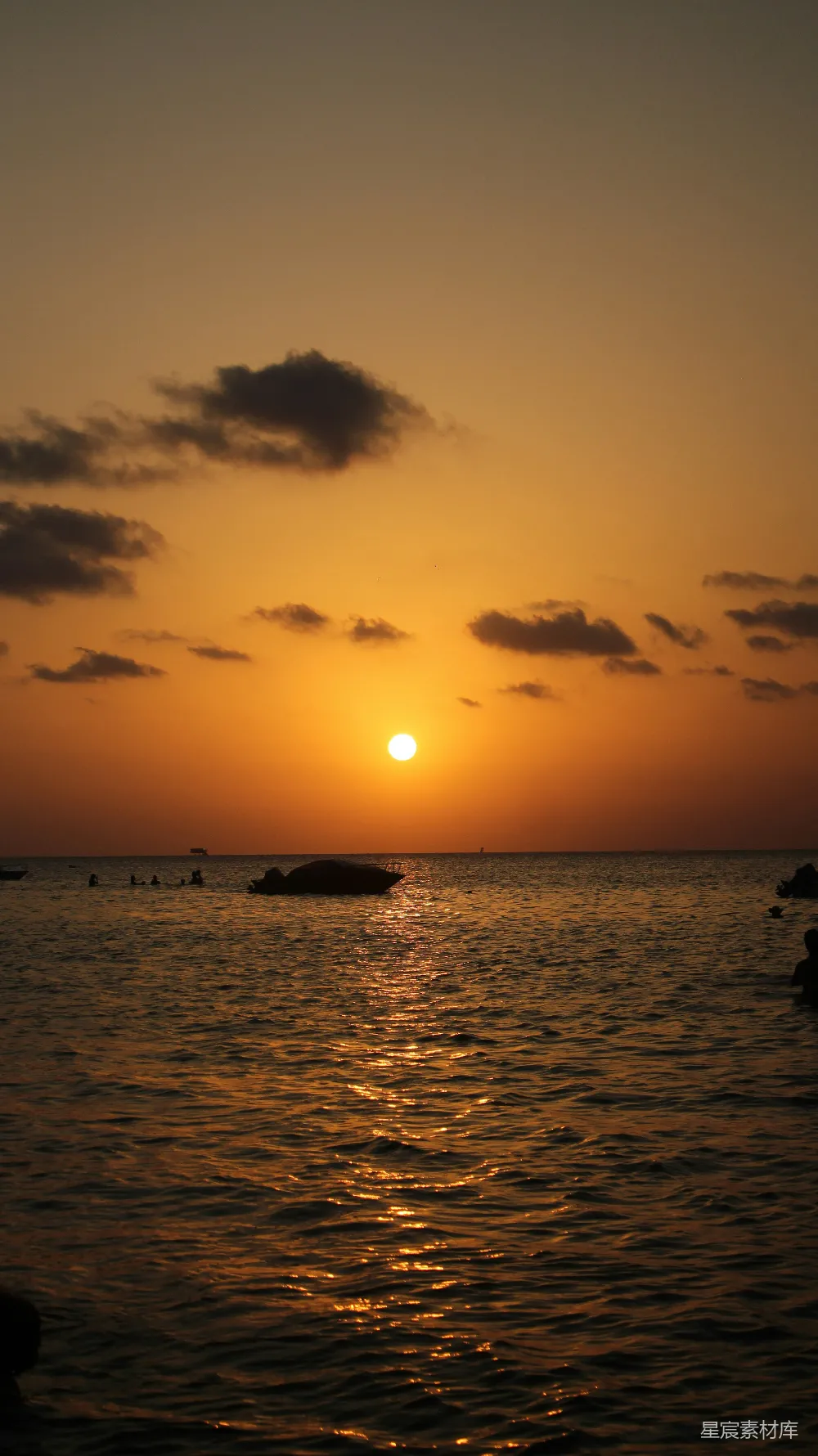 这张图片展示了一幅宁静的海景，夕阳洒在水面上，金色的光芒反射在波光粼粼的海面上，天空中有几朵云彩，远处有几个人在水中游泳，整体氛围非常平和，适合用来表达宁静与自然的主题。