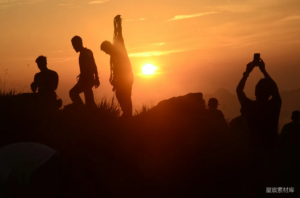 这张图片展示了夕阳下一群登山者在山顶的剪影。夕阳的余晖洒在他们的身上，形成鲜明的剪影效果，背景的山峰和天空也被染上了温暖的橙色。人们或坐或站，手中有的拿着绳索，有的举起手机拍摄，整个画面充满了探险和自由的气息。