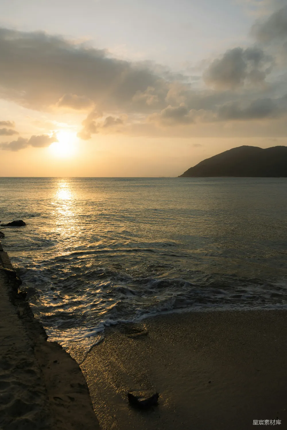 夕阳的余晖洒在平静的海面上，金色的光线与波浪交织，形成一幅宁静而美丽的画面。远处的山脉轮廓清晰，天空中散布着几朵云彩，增添了画面的层次感。这幅图适合用作旅行或自然风光的背景素材。