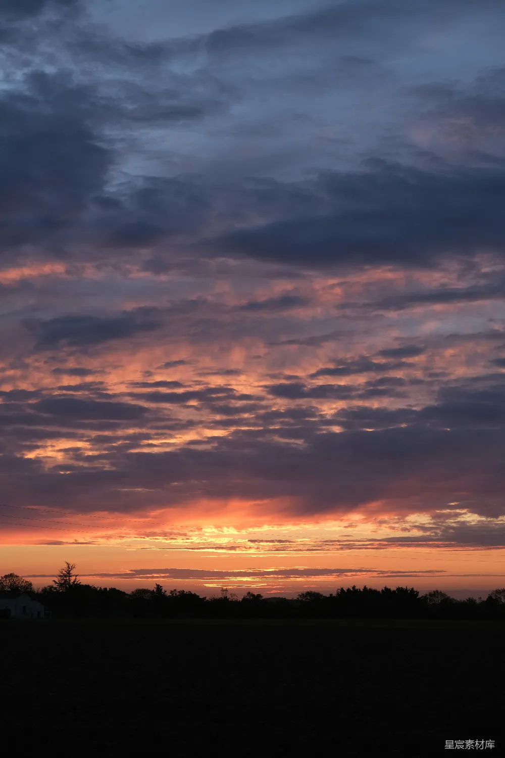 这张图片展示了一个宁静的田野，夕阳的余晖洒在云层上，天空被染成了橙红色，地平线处的树木和房屋在夕阳的光辉下显得格外宁静。适合用作自然风光的背景或壁纸。