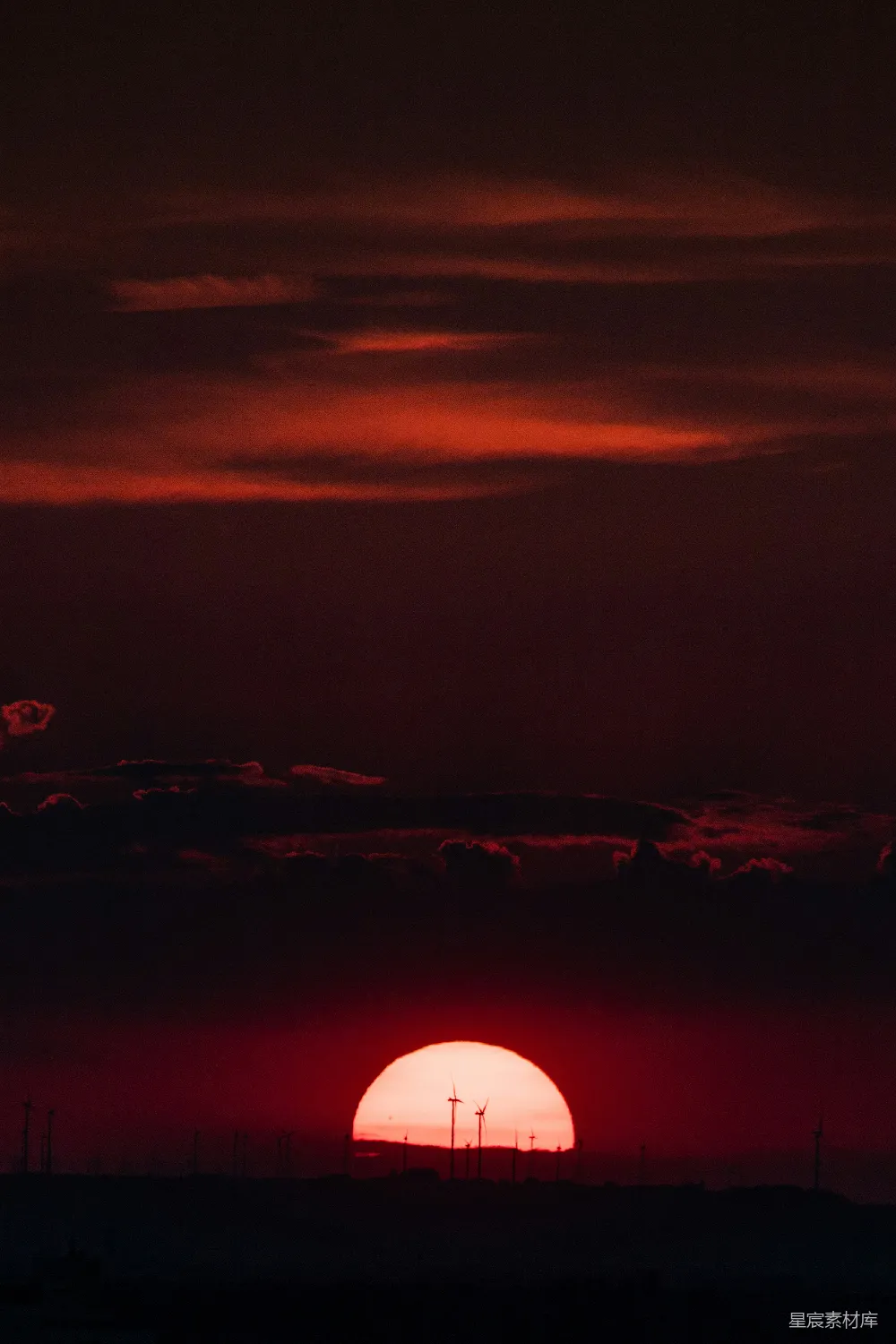 夕阳的余晖映照在风力发电站的叶片上，形成了一幅壮丽的工业与自然结合的风景。天空被染成了深红色，云层与地面的轮廓在夕阳的照射下显得格外清晰。这幅图展现了现代社会中可持续能源与自然美景的和谐共存。