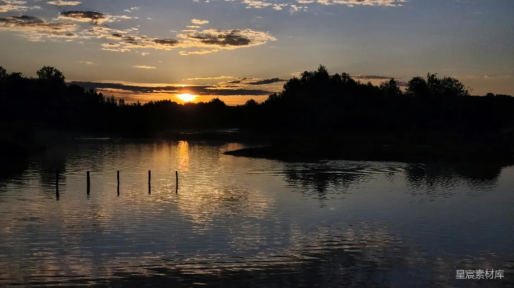 图片展示了一个宁静的湖面，夕阳的余晖洒在湖面上，形成美丽的倒影。湖面平静，只有几根木桩静静地漂浮。远处是茂密的树丛，与天空形成鲜明对比，营造出一种宁静而祥和的氛围。适合用于自然风光、放松心情或作为背景素材。
