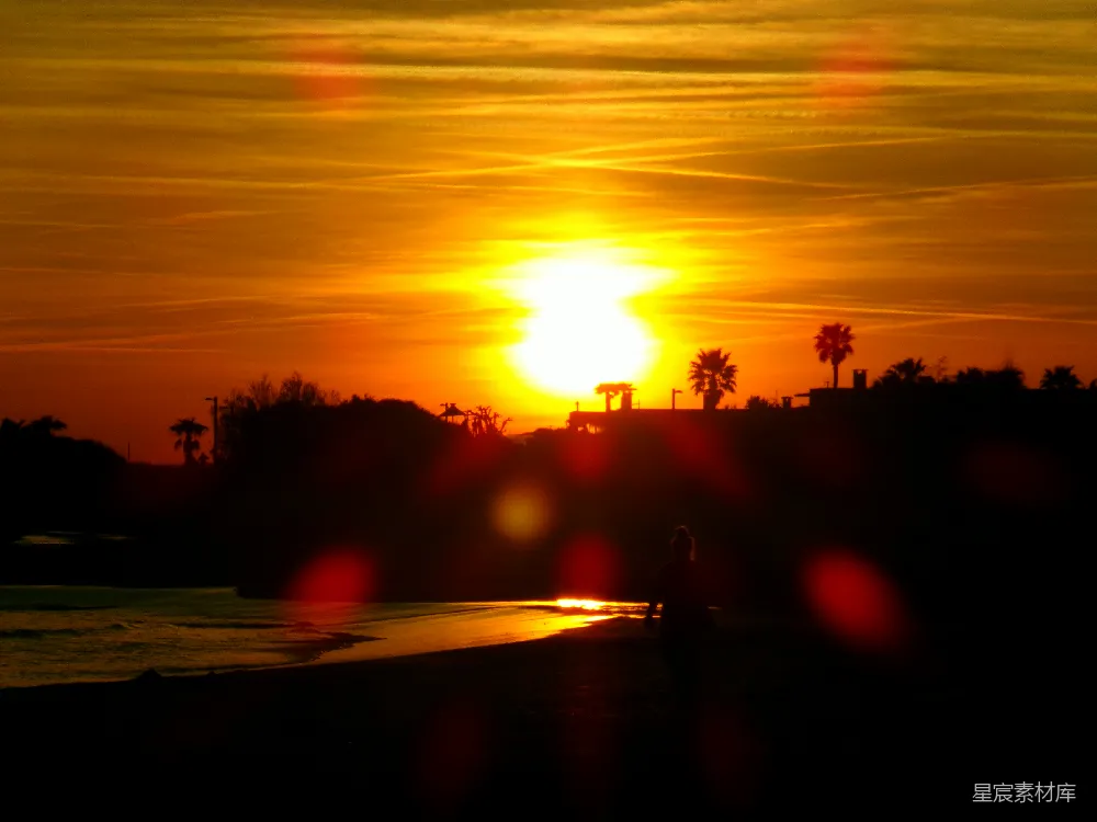 这张图片展示了一个黄昏时分，一个人在海滩上漫步的场景。夕阳的余晖洒在大地上，天空被染成了金黄色和橙色，远处的棕榈树和建筑剪影在落日的光辉中显得格外宁静。前景的光斑增添了一种梦幻的氛围，整体画面给人一种平和与放松的感觉。