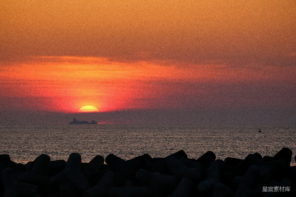 图片展示了一幅海港夕阳的美丽景象。夕阳将天空染成了橙红色，海面平静，远处有一艘船的轮廓清晰可见。前景的岩石结构为画面增添了层次感，营造出一种静谧而宁静的氛围。适合用于自然风光、旅游摄影或作为日落主题的背景素材。