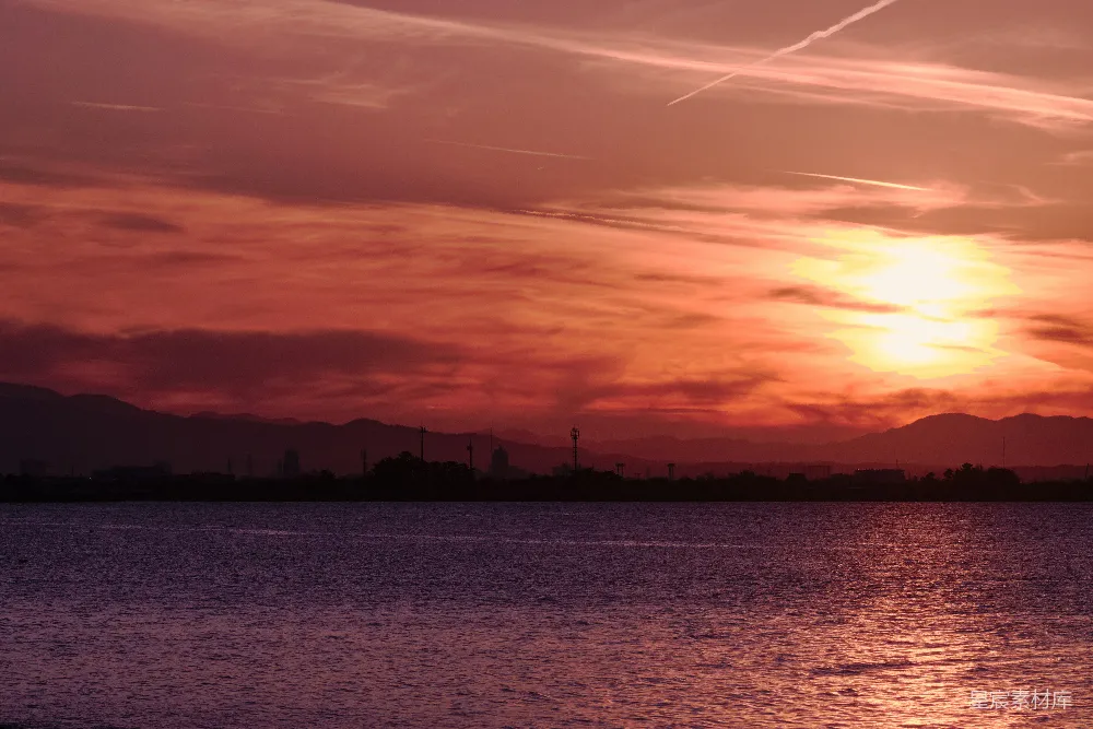 这张图片展示了日落时分的宁静湖景。夕阳的余晖洒在湖面上，天空呈现出橙红色的渐变，远处的山脉和城市的轮廓在黄昏的光线下显得格外柔和。湖水在夕阳的映照下波光粼粼，整个场景显得非常宁静和美丽。适合用作自然风光、旅行摄影或城市生活的背景图片。