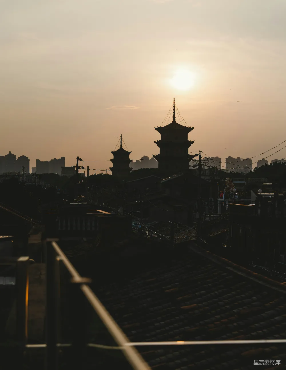 这张图片展示了夕阳下两座古塔的剪影，背景是城市的轮廓，天空呈现出柔和的橙黄色调。前景中的屋顶在夕阳的照射下显得有些昏暗，给人一种宁静而庄重的感觉。适合用作旅行、历史或自然风光的主题背景。