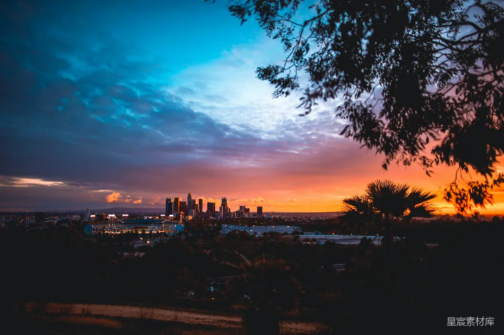 这张图片展示了洛杉矶的日落美景，城市的天际线在夕阳的余晖中显得格外壮丽，天空被染成了温暖的橙红色，树木和棕榈树的剪影增添了自然的元素。适合用作城市风光、旅游宣传、自然美景的素材。