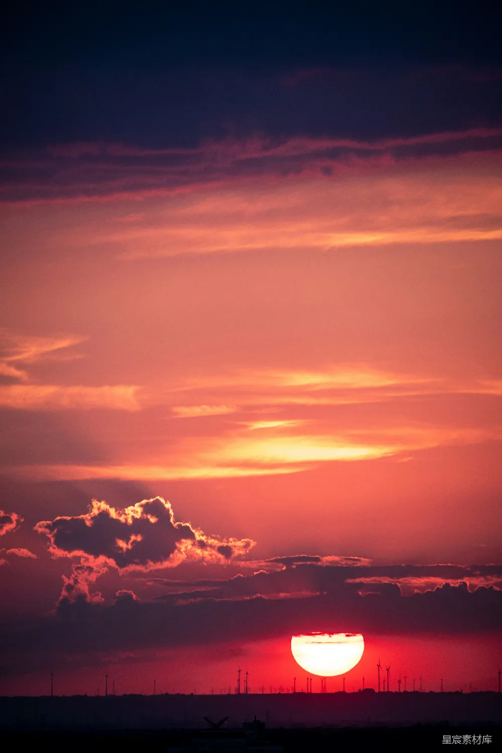 夕阳的余晖洒在地平线上，天空被染成了橙红色。地平线远处排列着一排排风车，它们在夕阳的映照下显得格外宁静而美丽。这幅画面捕捉了自然与人类文明的和谐共存，适合用作背景图片或手机壁纸。