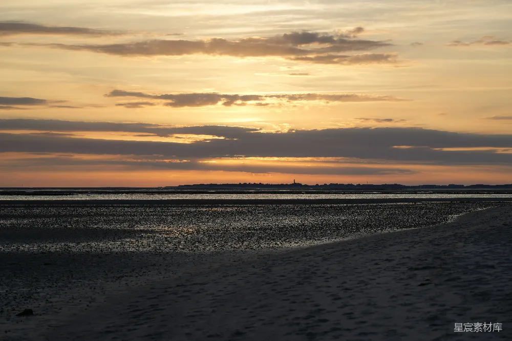 图片展示了一个宁静的海滩场景，夕阳的余晖洒在天空，云层被染成了金黄色。海面反射着夕阳的光芒，远处的城市轮廓在地平线上若隐若现，整个画面给人一种平静而祥和的感觉。适合用于展现自然之美、旅行和放松的主题。