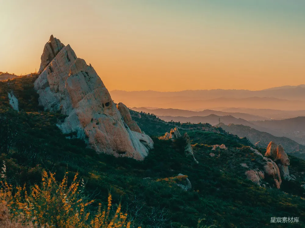 这张图片展示了一处壮丽的日落场景，夕阳的余晖洒在崎岖的岩石上，山峦在柔和的光线中若隐若现。前景的黄色小花为画面增添了生机，呈现出一种宁静而壮丽的自然风光。适合用于自然风光、户外旅行或日落主题的背景。
