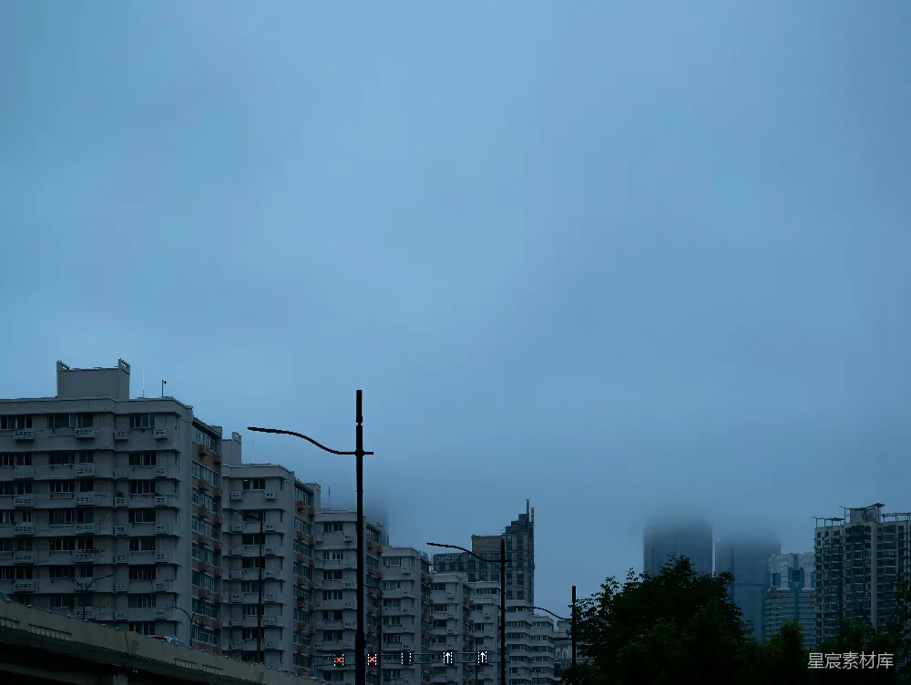 这张图片展示了一个清晨或傍晚的城市景象，天空中弥漫着淡淡的雾气，远处的高楼大厦若隐若现，近处的建筑清晰可见。路灯和交通信号灯在雾中显得格外醒目，营造出一种宁静而神秘的氛围。适合用于城市风光、雾景、清晨等主题的背景图。