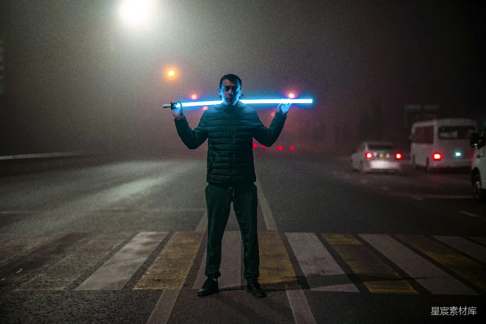 这张图片展示了一位男子在雾蒙蒙的街道上手持一根蓝色光剑，背景是城市的夜景和模糊的车辆灯光。男子站在斑马线上，整体画面充满了神秘和科幻的氛围，适合用于展示夜晚城市中的独特风景和创意摄影。