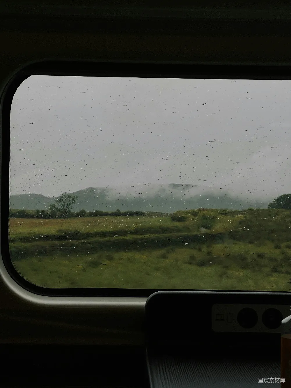这张图片展示了一列火车内部的景象，透过窗户可以看到外面是一片雨中的风景。远处的山脉在云雾中若隐若现，而近处则是一片郁郁葱葱的草地，给人一种宁静而深邃的感觉。雨滴在窗户上留下了痕迹，增添了一份独特的氛围。这张图片适合用作旅行、自然和宁静主题的