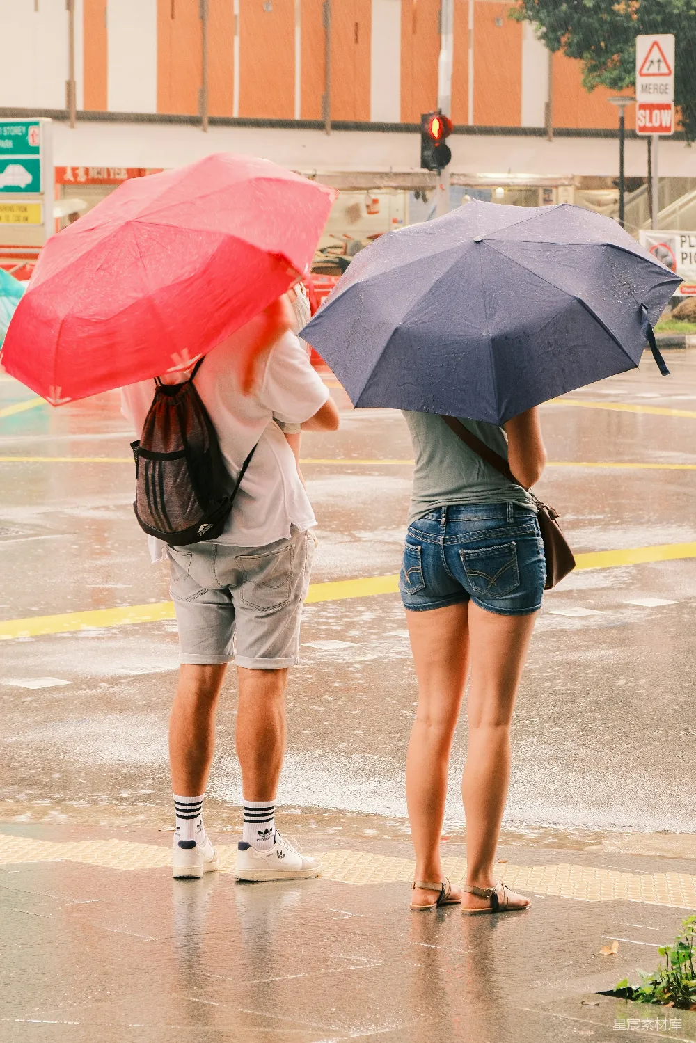 这张照片展示了两位在雨中并肩而立的情侣，他们各自撑着颜色鲜艳的雨伞，穿着适合夏季的短裤和T恤，背景是城市街道和交通信号灯。雨滴在伞面上闪闪发光，营造出一种温馨而浪漫的氛围。适合用作情侣故事、城市风情或情感主题的背景素材。