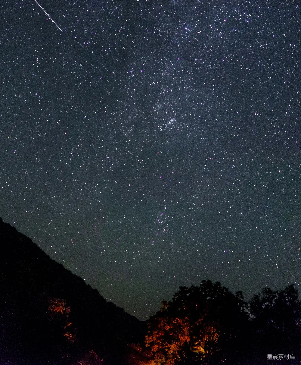 这张图片展示了夜空中无数星星的璀璨景象，流星划过天际，为宁静的夜空增添了一抹动感。山峦与树木在黑暗中若隐若现，与星空形成鲜明对比，营造出一种神秘而宁静的氛围。适合用于表达对自然美景的赞美，或者用于制作夜景主题的背景素材。