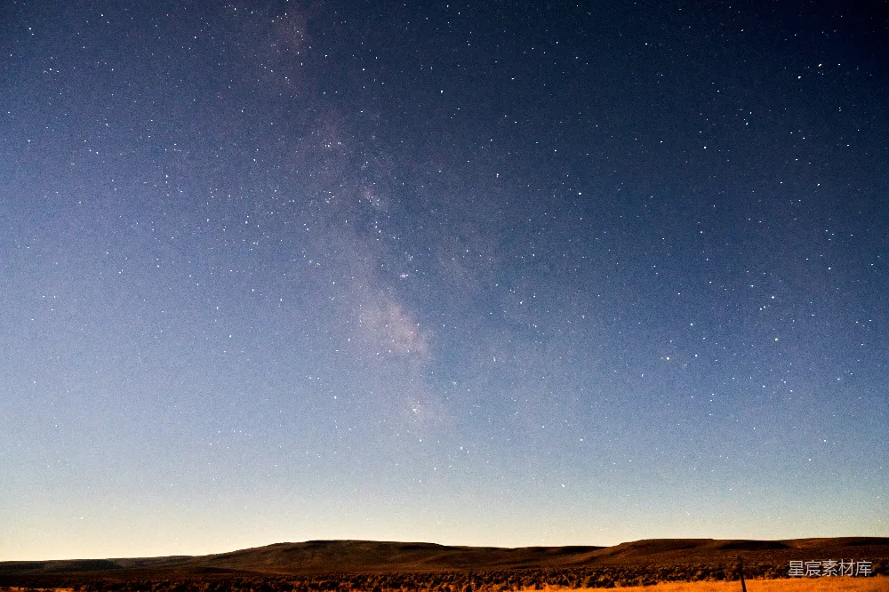 这张图片展示了一个宁静的夜景，天空中星星点点，银河清晰可见，地平线上的山丘和树木勾勒出一幅宁静的自然景象。整体色调柔和，给人一种宁静和深邃的感觉，适合用于自然风光、宁静生活或夜空主题的背景。