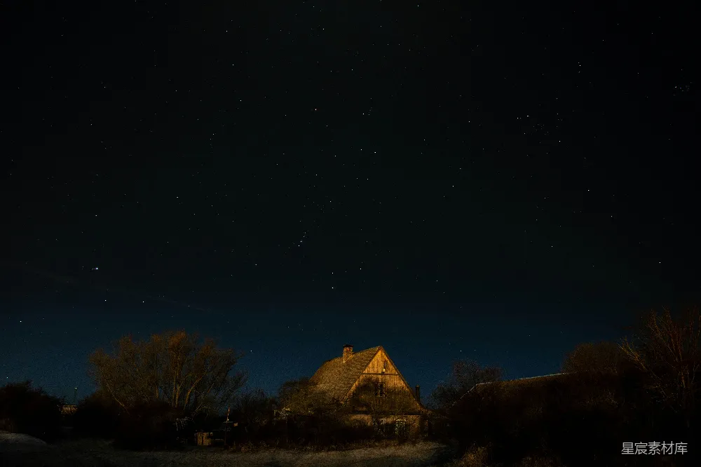 这张图片展示了一座位于乡村的小屋在夜空中被璀璨的星星照亮的景象。天空深邃，星星闪烁，小屋在微弱的灯光下显得格外温馨。周围的树木在黑暗中若隐若现，营造出一种宁静而神秘的氛围。这幅图片适合用于描述乡村夜景、自然摄影或静谧的乡村生活。