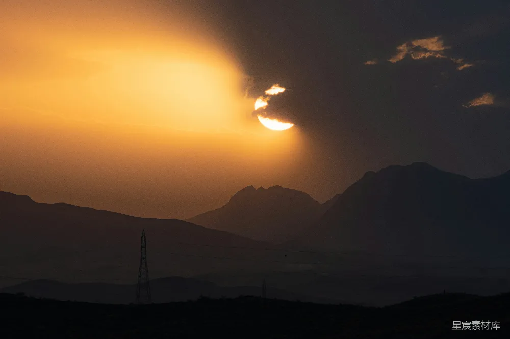 图片展示了一幅夕阳下的山脉景象，夕阳的光芒透过云层洒在山脉上，形成了美丽的剪影效果。天空中云朵轻柔，地面上的山脉轮廓清晰，远处还有一座电线塔。整个画面宁静而壮丽，适合用作自然风光的背景或创作灵感。