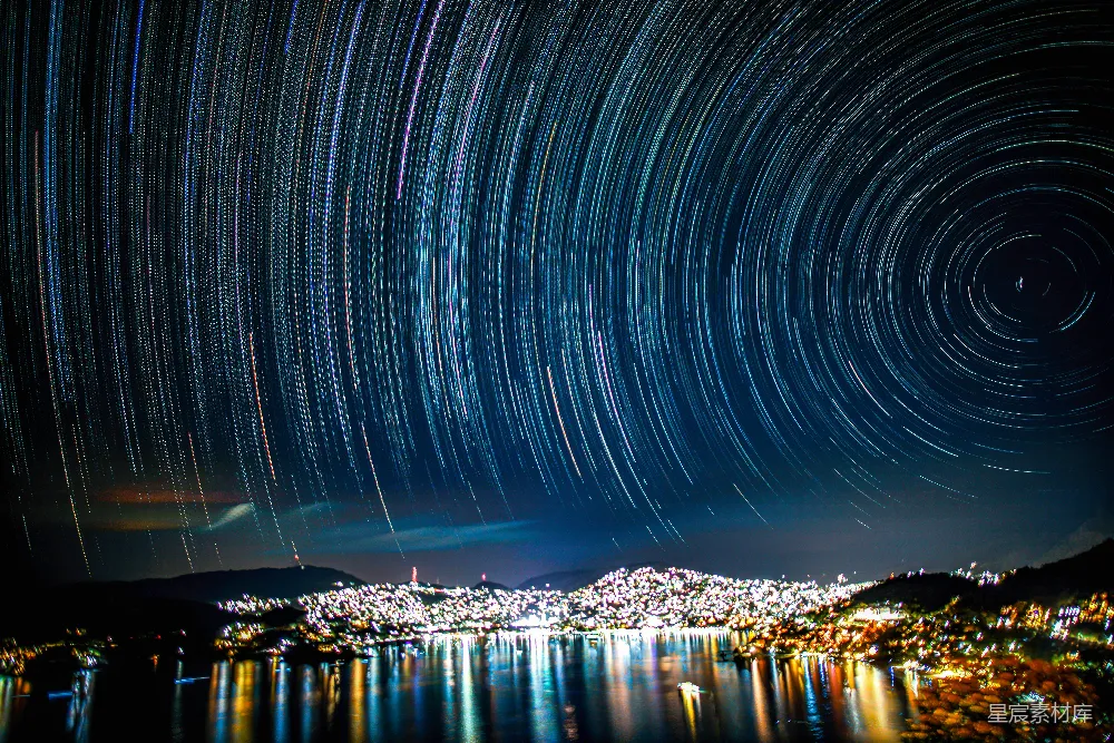 这张图片展示了星轨现象下的城市夜景。星轨的旋转轨迹与城市灯光交相辉映，形成了一幅壮丽的夜景。城市的灯光在水面上倒影，增添了画面的层次感和美感。这张图片适合用作自然风光、城市夜景或摄影爱好者的作品展示。