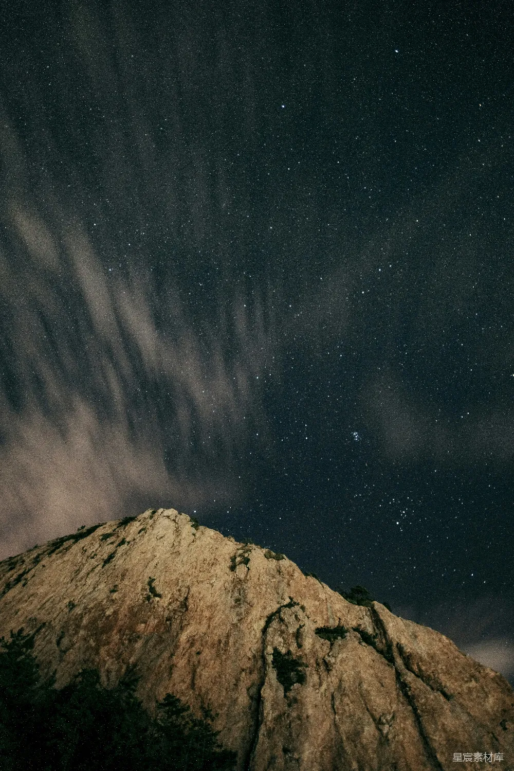 这张图片展示了夜晚山峰的壮丽景色，山峰的轮廓在星光的映衬下显得格外清晰。夜空中布满了星星，银河横跨天际，云层在夜空中缓缓移动，营造出一种宁静而神秘的氛围。适合用于自然风光、星空摄影或静谧夜景的主题。