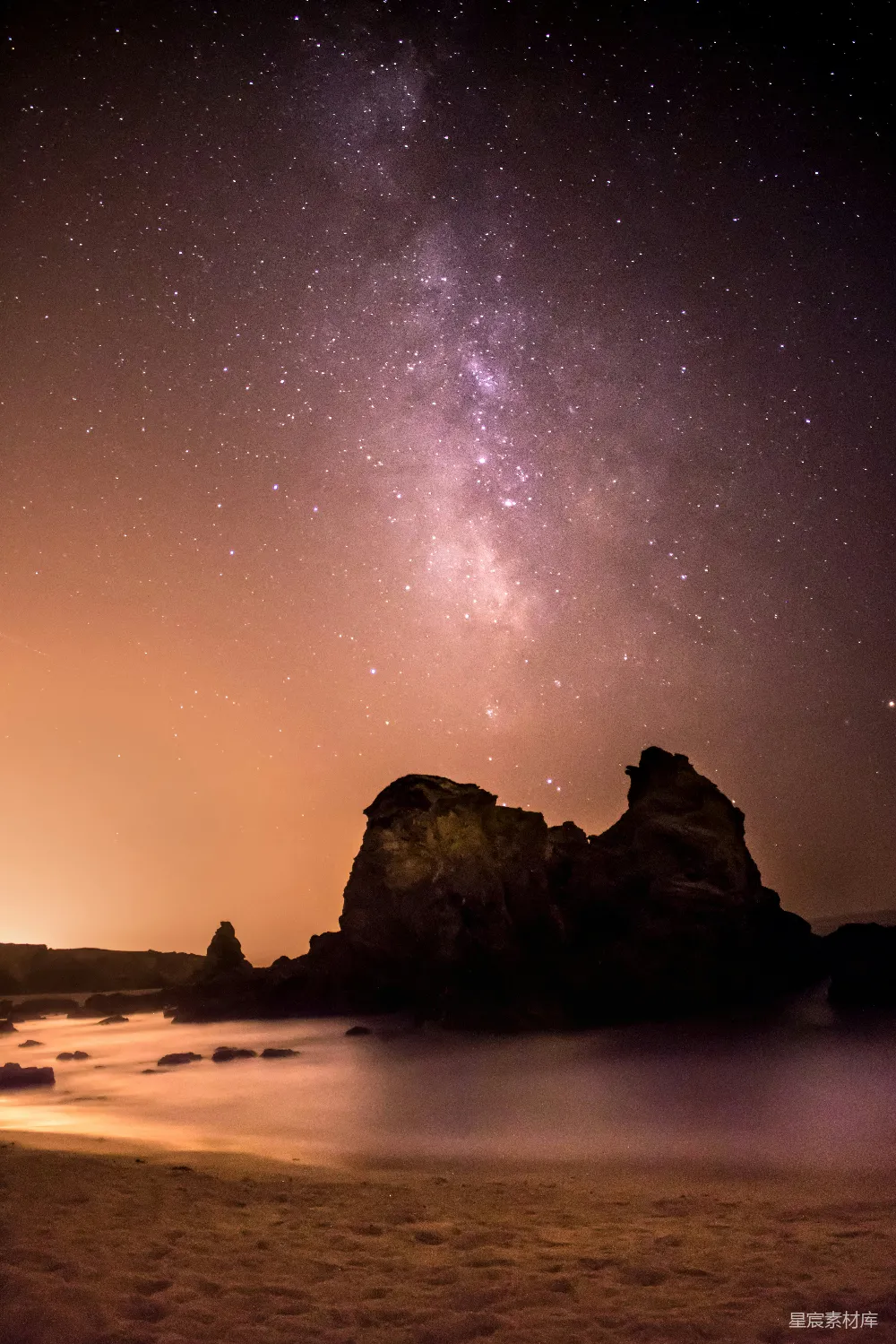 这张图片展示了一片岩石海岸在夜晚的景象，天空中布满了星星和银河，地平线附近有一座岩石，海水轻轻拍打着岩石，营造出宁静而神秘的氛围。适合用于展示自然之美、星空摄影以及夜景摄影等场景。
