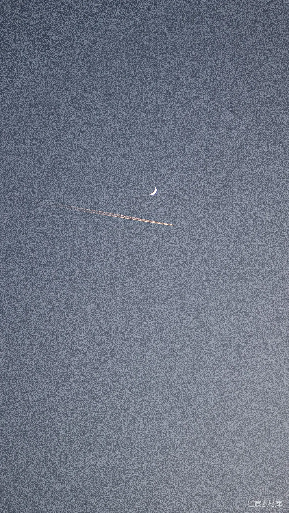 这张图片展示了一个宁静的夜空场景，新月在夜空中显得格外明亮，而一架飞机的尾迹横穿画面，留下了长长的轨迹。整体色调偏冷，营造出一种宁静而神秘的氛围。适合用作手机壁纸或装饰画的背景。