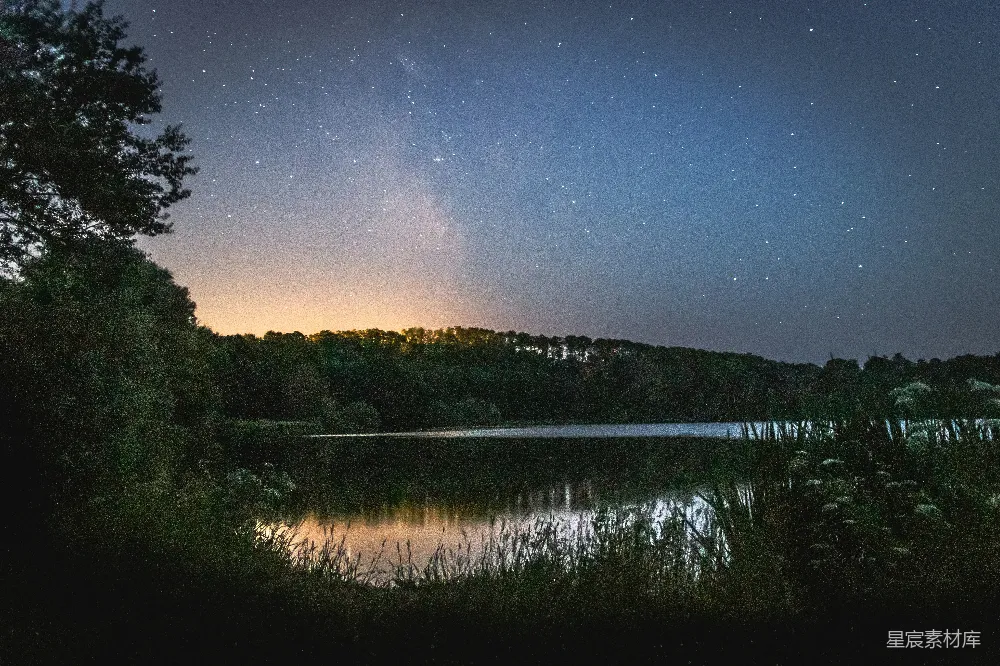 这张图片展示了一个宁静的湖畔夜景，湖面倒映着树木和天空中的星辰。天空中布满了繁星，银河清晰可见，营造出一种宁静而神秘的氛围。适合用作自然风光、旅行摄影或静谧夜景的背景素材。