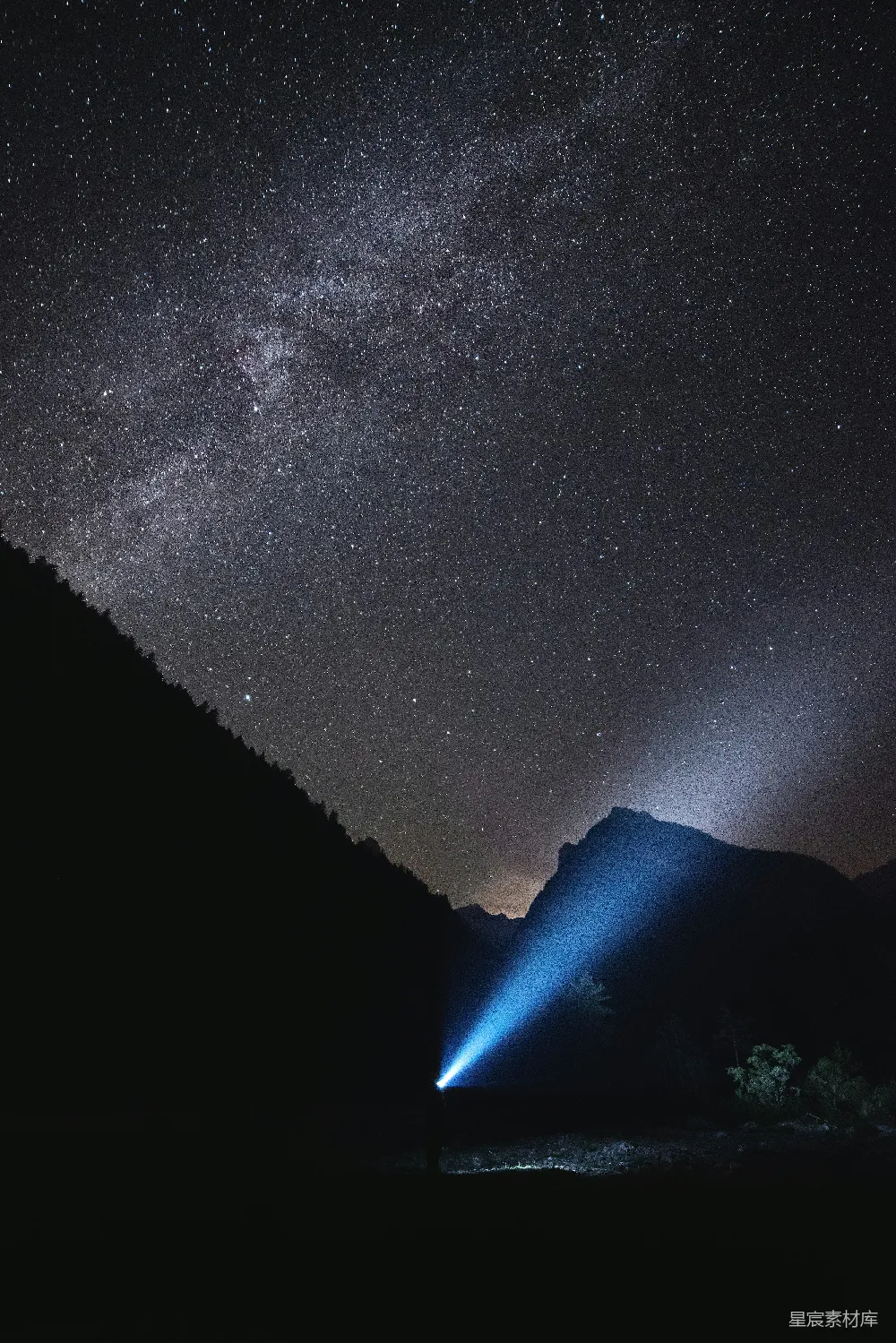一张夜空下的照片，银河横跨夜空，明亮的手电筒光束从山脚下射出，照亮了前方的黑暗，营造出一种宁静而神秘的氛围。