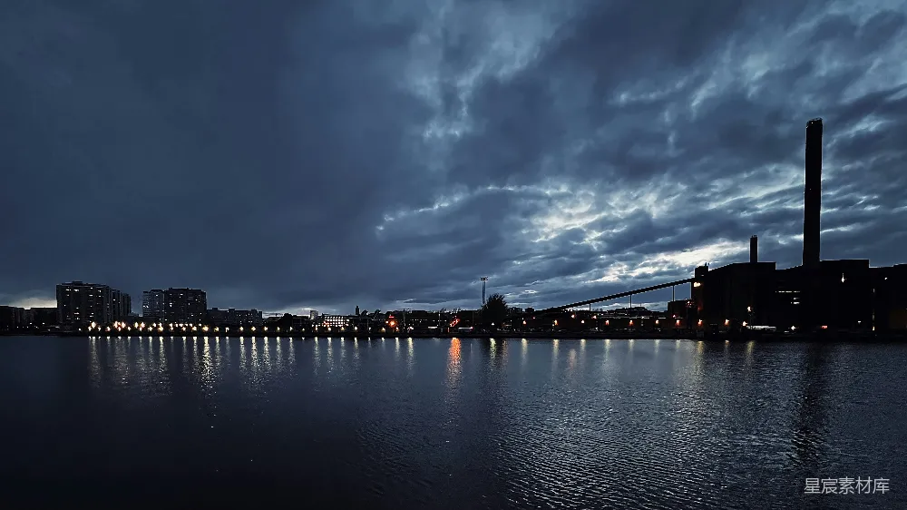 这张图片展示了夜幕下宁静的城市湖景。深邃的天空中布满了厚重的云层，湖面平静如镜，倒映着城市的灯光和建筑的轮廓。远处的高楼灯火辉煌，近处的烟囱和桥梁在夜色中显得格外突出。整个场景显得既宁静又神秘。
