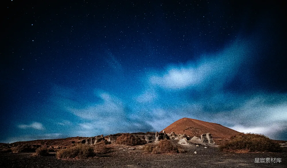 这张图片展示了一座火山在夜空下的景象，星空点缀其中，云层轻柔地飘过，地面上的植被稀疏，整体氛围宁静而神秘。适合用于自然风光、摄影爱好者或宁静主题的背景素材。