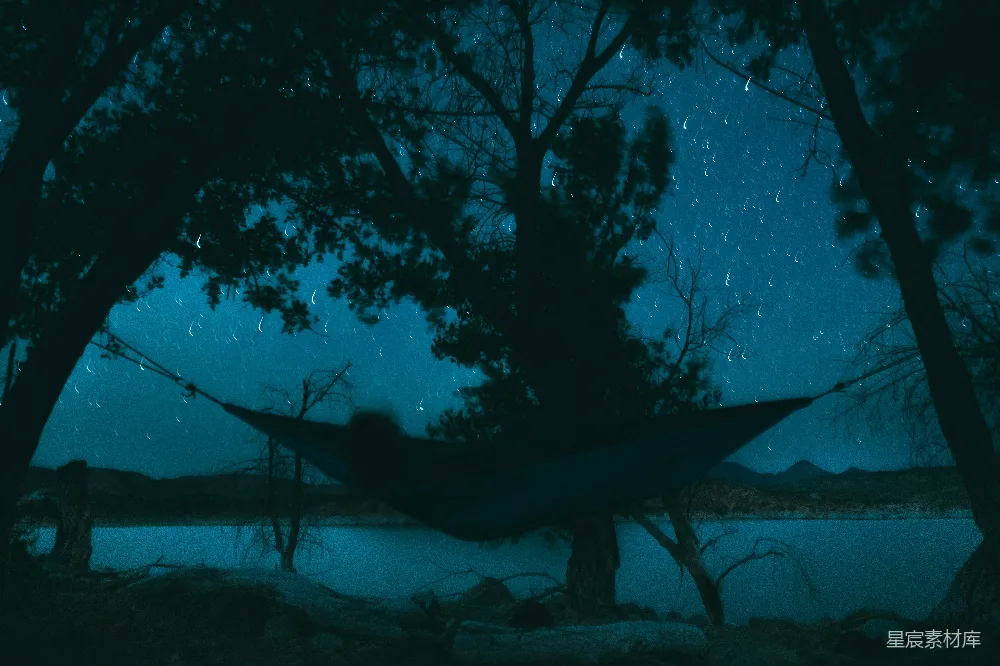 这张图片展示了一个宁静的夜晚场景，吊床悬挂在两棵树之间，背景是星空和远处的山脉。深蓝色的色调和稀疏的树木营造出一种宁静而孤独的氛围，适合用来表达户外旅行、自然探索或夜晚放松的主题。