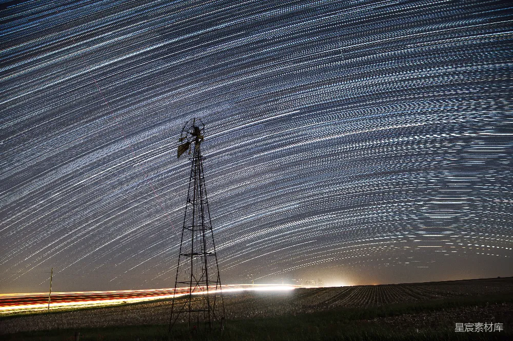 这张图片展示了一幅宁静的乡村夜景，画面中央有一座风车，背景是夜空中清晰可见的星轨，仿佛银河倾泻而下。地平线处有微弱的灯光，增添了画面的层次感。整幅画面既有自然的宁静，又有科技的痕迹，适合用于展现自然美景与现代科技结合的主题。