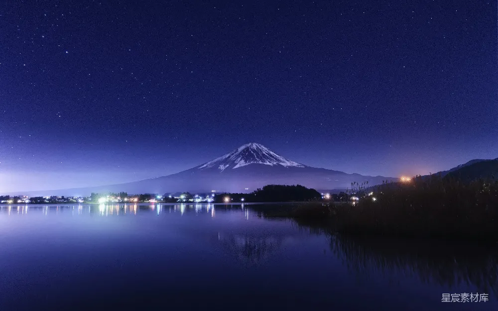 这张图片展示了夜晚时分，富士山在星空下的壮丽景色。远处的富士山被蓝色的灯光照亮，倒映在平静的湖面上，形成美丽的星空倒影。背景中的夜空星星点点，营造出宁静而神秘的氛围。这张图片适合用作旅行、自然风光或夜景的背景素材。