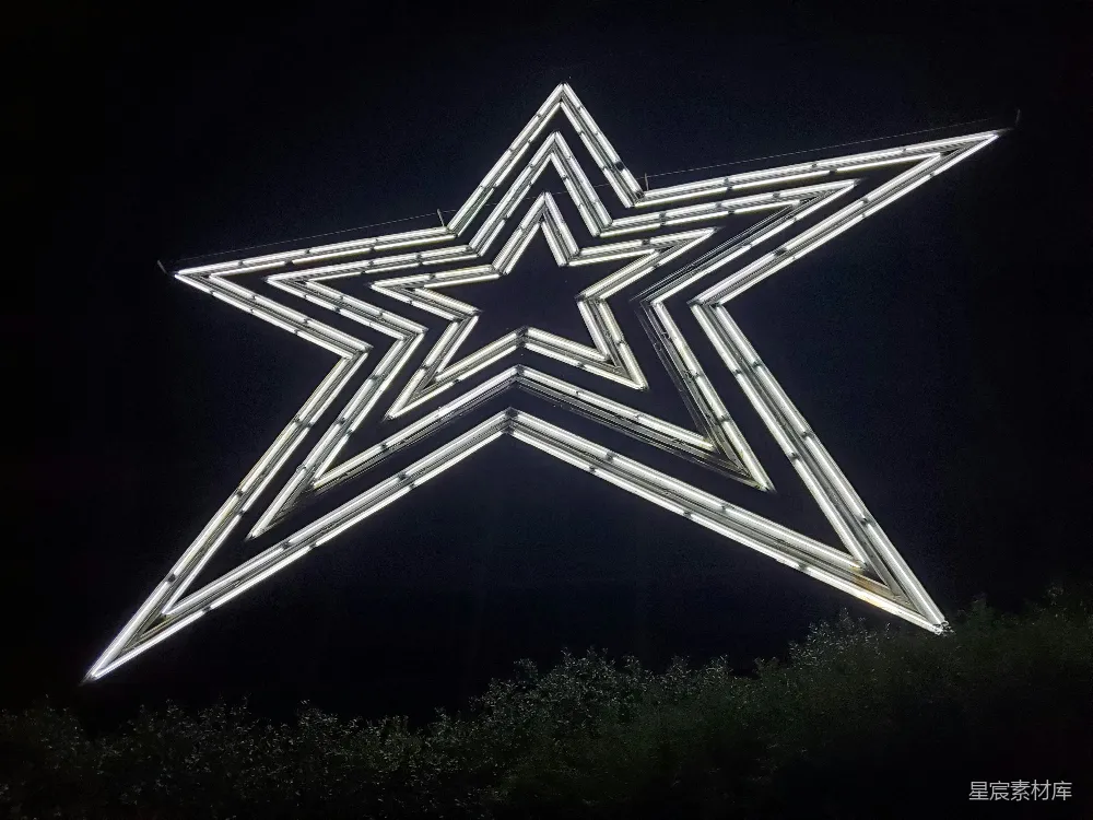 这张图片展示了一个巨大的五角星灯饰，夜晚被点亮，发出耀眼的光芒。五角星由许多细长的灯管组成，形成了一个简洁而现代的几何形状。这个灯饰悬挂在空中，下方是模糊的树木轮廓，营造出一种梦幻的氛围。适合用于节日庆典、夜间娱乐活动或城市景观照明。