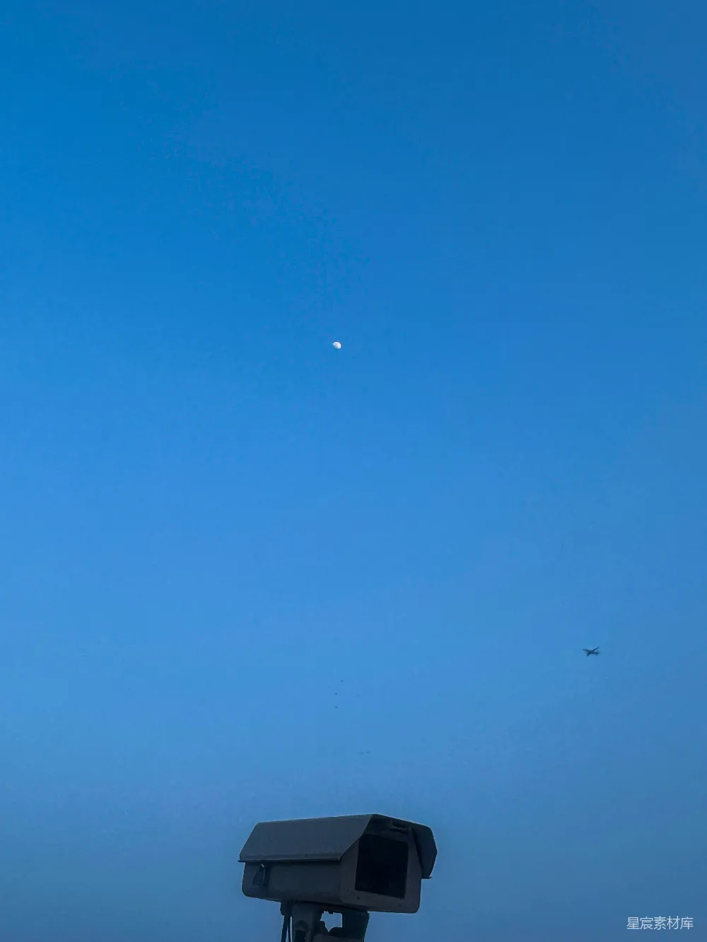 这张图片展示了一个宁静的蓝天背景，画面中央有一架飞机在飞行，下方是一个监控摄像头。背景的蓝天上有一个小的月亮，整个画面给人一种宁静而自然的感觉，适合用于表达平和与观察的主题。